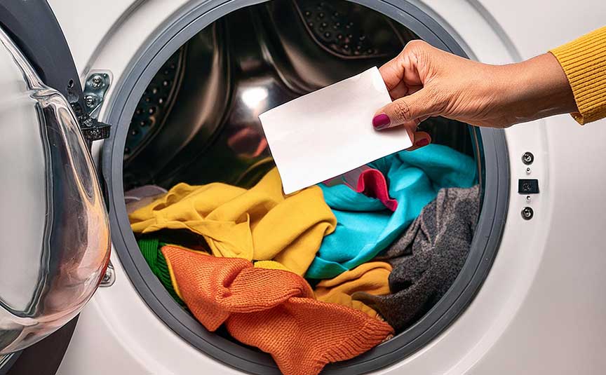 Firefly colorfull clothes that are placed in a washing machine. A hand is holding a laundry detergent sheet in front.